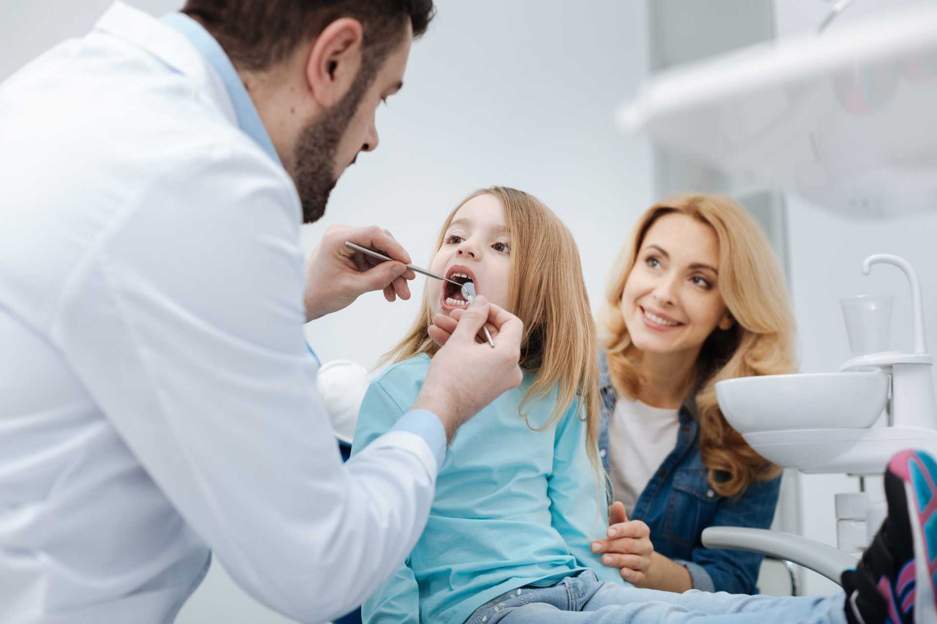 Attentive,mom,brining,her,child,to,the,dentist Woman sitting behind girl with mouth open and Dr inspecting teeth