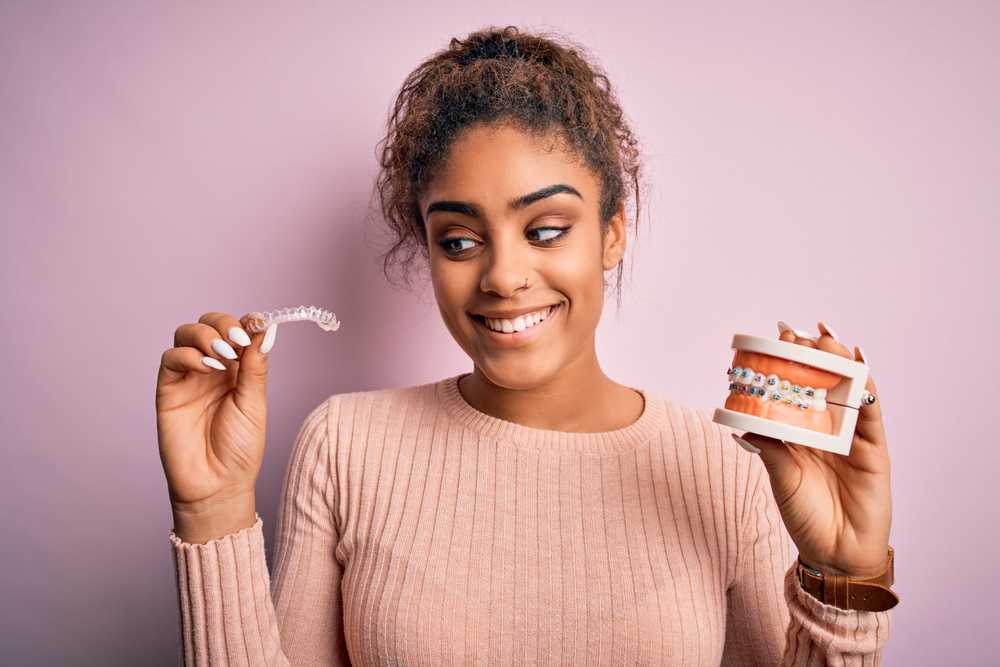 Young,african,american,woman,smiling,happy,holding,professional,orthodontic,denture Woman holding model of teeth with braces in one hand and invisalign retainer in the other